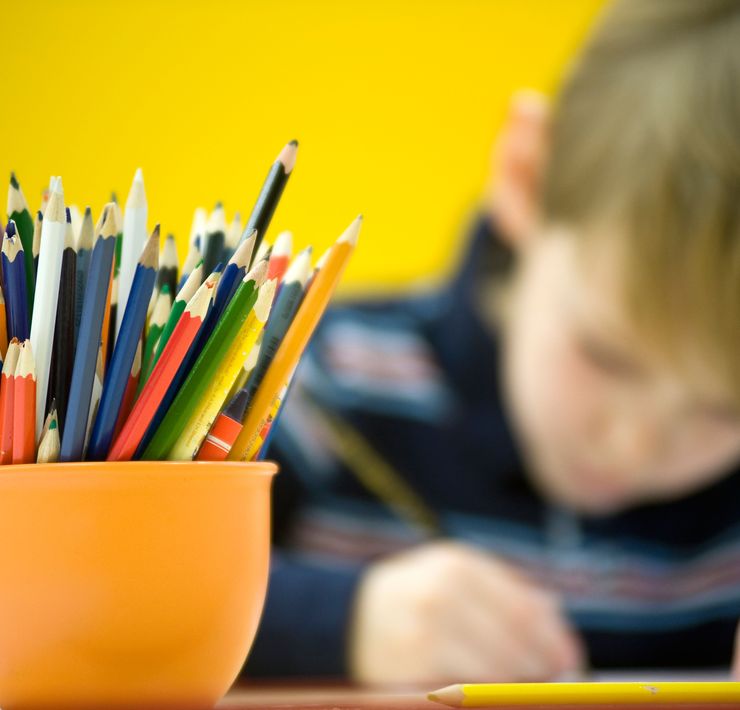 Ein Kind sitzt an einem Tisch und hält einen Stift in der Hand. Im Vordergrund steht ein Gefäß mit vielen, dünnen Buntstiften