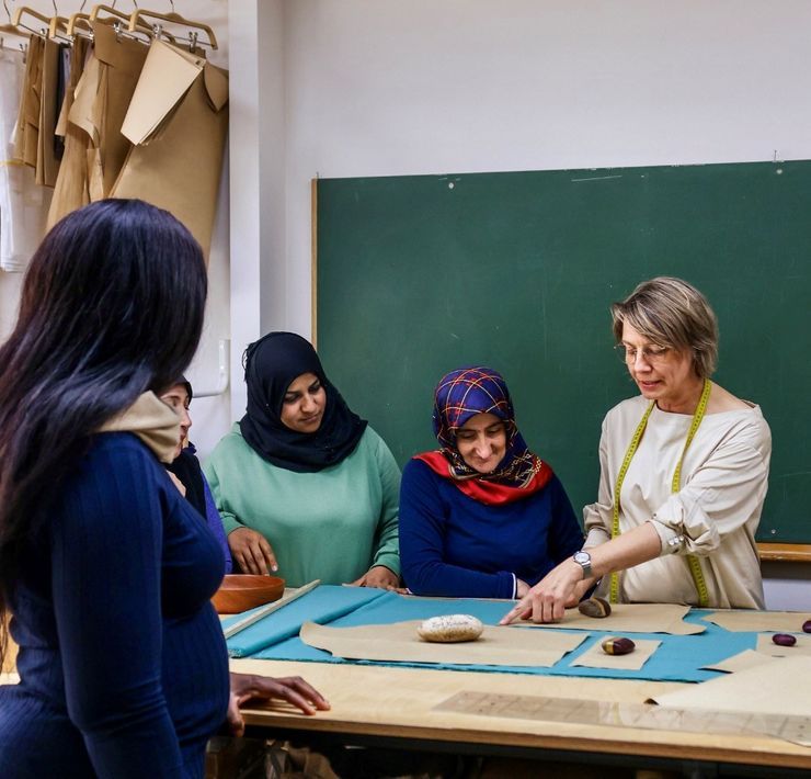 Eine Frau mit Brille steht an einem Tisch und deutet auf Schnittmuster, die mit Steinen befestigt auf einer türkisfarbigen Stoffbahn liegen. Um sie herum stehen mehrere Frauen, mutmaßlich mit Migrationshintergrund, die ihr zuhören.