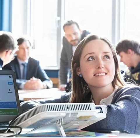 Eine junge Frau in einem Klassenzimmer hockt neben einem Beamer, der mit einem Laptop verbunden ist und ein Bild an die Wand wirft. Im Hintergrund sind weitere Personen an Tischen zu sehen.
