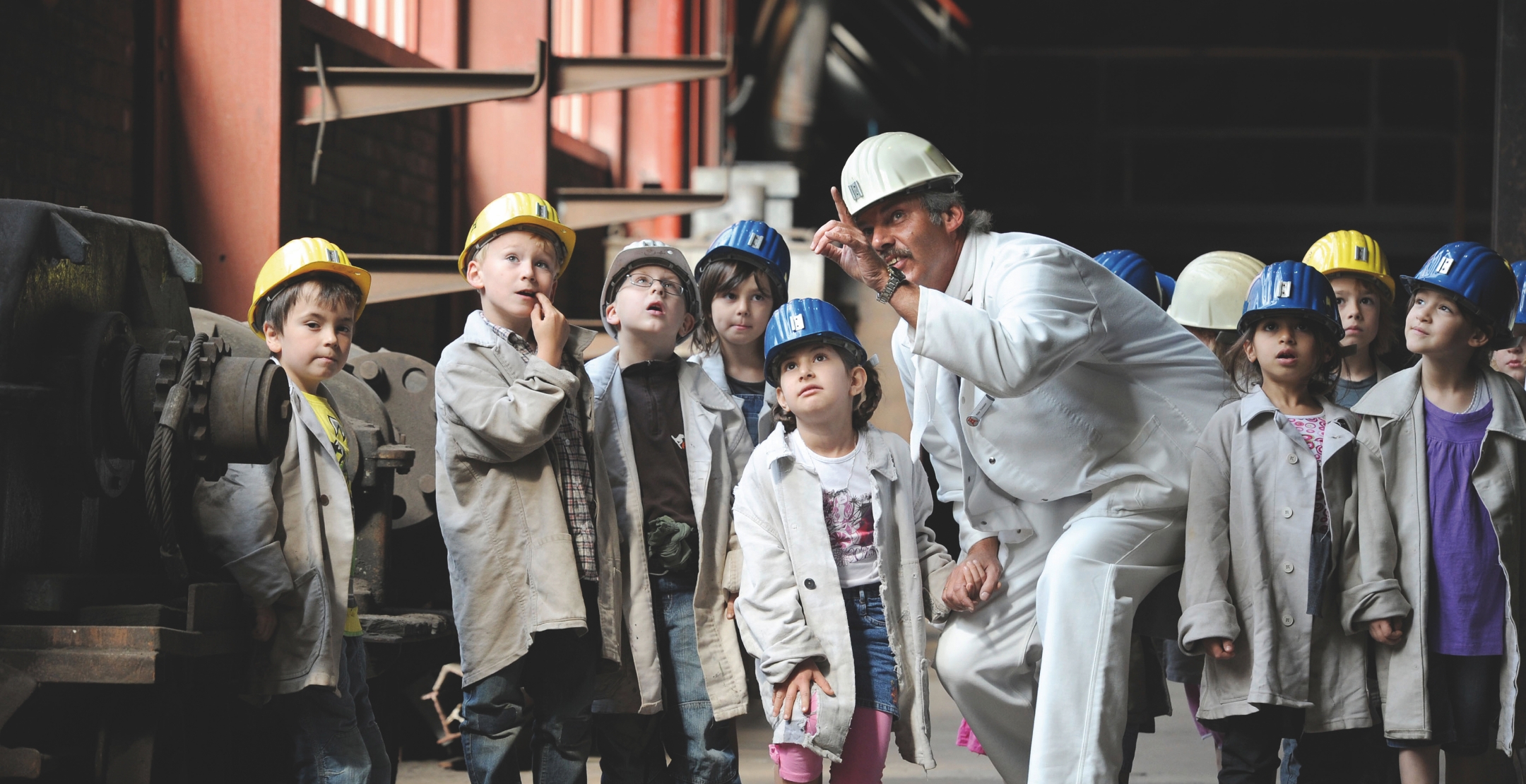 Ein Mann mit Bergmannskittel und Helm zeigt staunenden Kindern im Grundschulalter etwas in den Räumlichkeiten der Zeche Zollverein. Die Kinder tragen bunte und gelbe Kinderhelme und ebenso weiße Bergmannskittel.