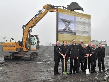 Spatenstich auf der Halde durch verschiedene Projektbeteiligte in schwarzer Kleidung. Im Hintergrund ein gelber Bagger mit einem Foto der Visualisierung. Der Himmel ist grau, der Boden schlammig.