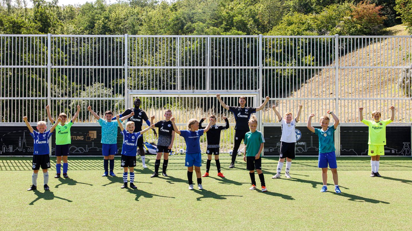 Eine Gruppe bestehend aus Kindern und Trainern, mutmaßlich auch mit Migrationshintergrund, die auf einem eingezäunten Bolzplatz stehend ihre Hände nach oben strecken und jubeln. 