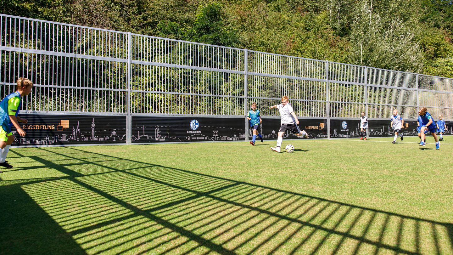 Kinder spielen Fußball auf dem Rasen des eingezäunten Bolzplatzes. An der Bande sind die Logos von Schalke hilft! und der RAG-Stiftung zu sehen.