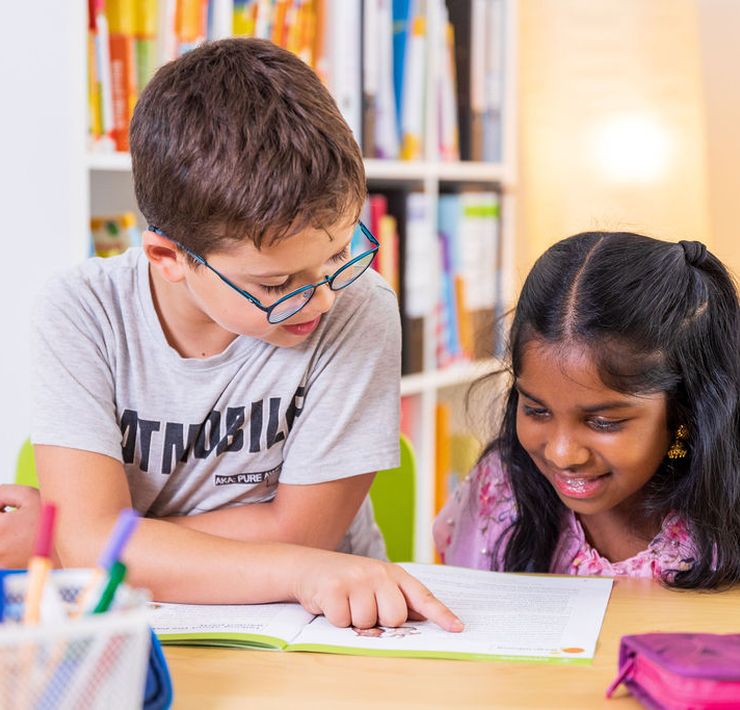 Ein älteres Kind mit Brille erklärt einem jüngeren Kind etwas in einem Lehrbuch. Beide sitzen dazu an einem Tisch, auf dem auch Stifte und ein Etui bereitstehen.