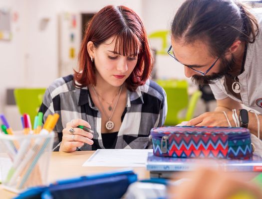 Ein Studierender mit Bart und Brille erklärt einer Jugendlichen etwas auf einem Blatt Papier. Etui und Stifte stehen bereit.