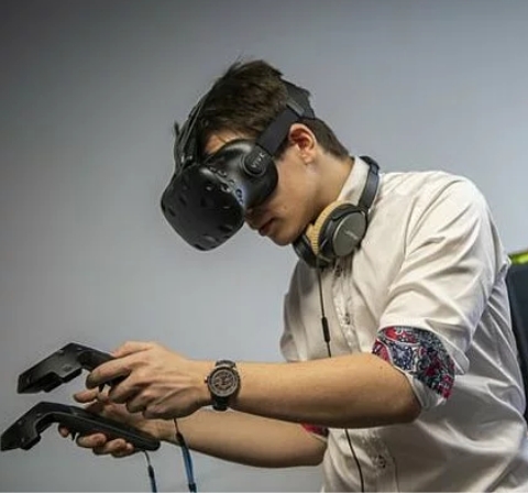 Ein Jugendlicher hält zwei Steuerungselemente in der Hand und trägt dabei eine VR-Brille. Ein Bügelkopfhörer hängt um seinen Hals.