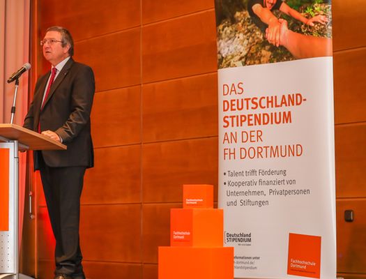 Ein Mann steht an einem Rednerpult und spricht in ein Mikrofon. Im Hintergrund ist ein Roll-up zu sehen. Auf diesem steht unter anderem "Das Deutschlandstipendium an der FH Dortmund"
