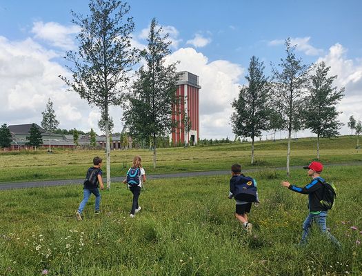 Vier Kinder mit Rucksäcken laufen über eine grüne Wiese. Im Hintergrund ist der Förderturm der Zeche Friedrich Heinrich zu sehen.