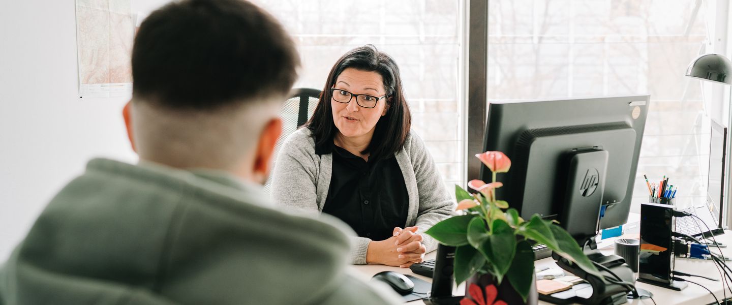 Eine Frau mit Brille sitzt an einem Tisch vor einem PC Monitor, auf dem auch Stifte und eine Blumenvase zu sehen ist. Vor ihr sitzt eine jüngere Person und hört zu.