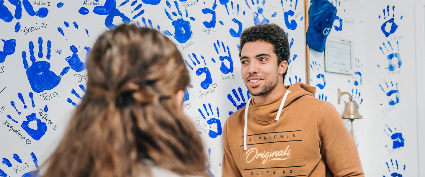 Ein junger Mann steht vor einer Wand mit vielen blauem Handabdrücken und Namen darunter. Vor ihm steht eine Person mit längeren welligen Haaren, die ebenso die Wand betrachtet.