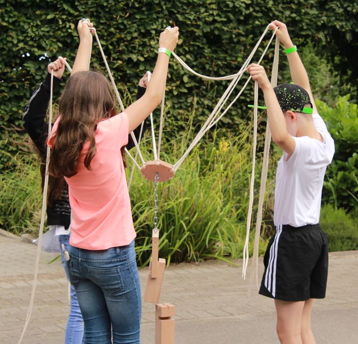 Drei Kinder einer weiterführenden Schule halten gemeinsam eine Seilkonstruktion in die Höhe, mit der sie versuchen ein Holzstück auf einem anderen Holzstück zu platzieren.