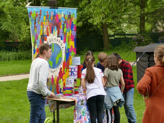 Eine Gruppe Jugendlicher steht vor einem Tisch mit bunt angemalten Dosen wie beim Dosenwerfen. Dahinter hängt ein buntes Plakat mit vielen farbigen Silhouetten von Menschen. Auf dem Plakat ist der Schriftzug "Jahrmarkt" zu lesen..