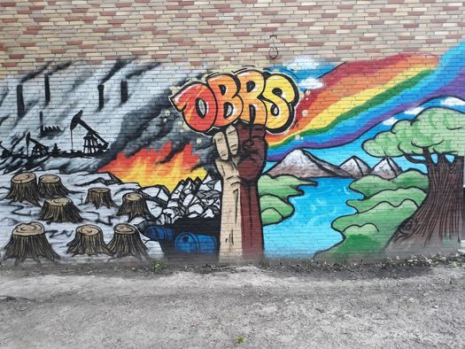 Ein Graffiti in dem links abgeholzte Baumstümpfe, umgestürzte Ölfässer und Silhouetten von Kränen des Tagebaus zu sehen sind. In der Mitte ist eine gestreckte Faust mit dem Schriftzug "OBRS" platziert. Rechts davon ist ein Regenbogen, ein Fluss, ein Berg sowie ein gesunder Baum zu sehen. 