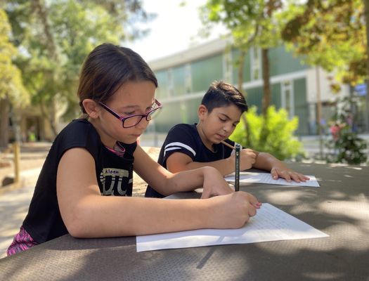 Zwei Kinder sitzen im Freien an einem Tisch. Sie haben Stifte in der Hand und schreiben konzentriert etwas auf Papier.