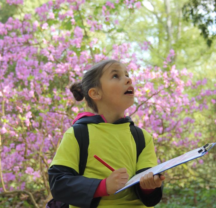 Ein Mädchen vor einem pink blühenden Strauch betrachtet die Natur. Sie hat dabei einen Notizblock und einen Stift in der Hand.