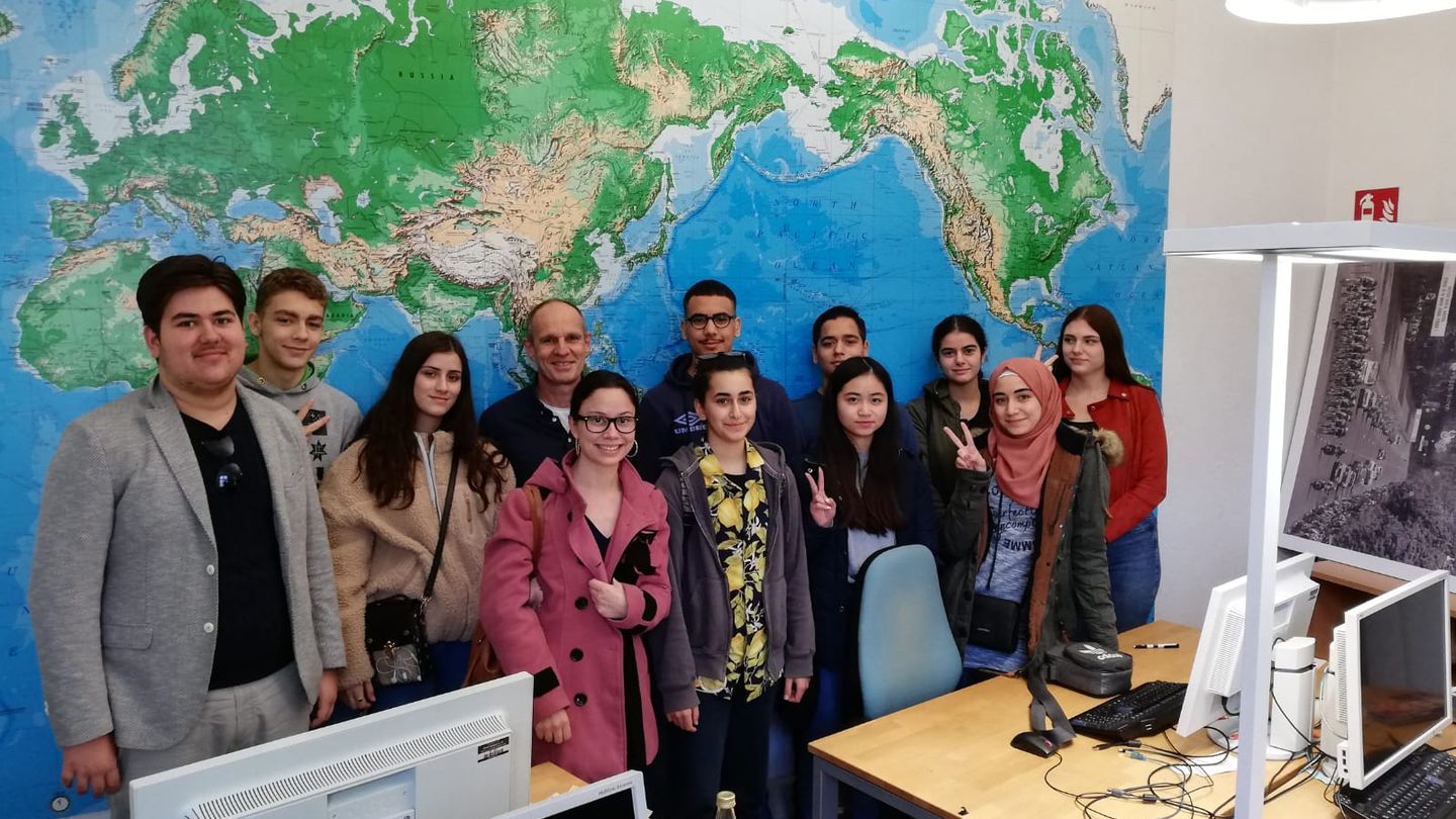 Eine Gruppe Jugendlicher, zum Teil mutmaßlich mit Migrationshintergrund, steht vor einer Weltkarte in einem Büro.