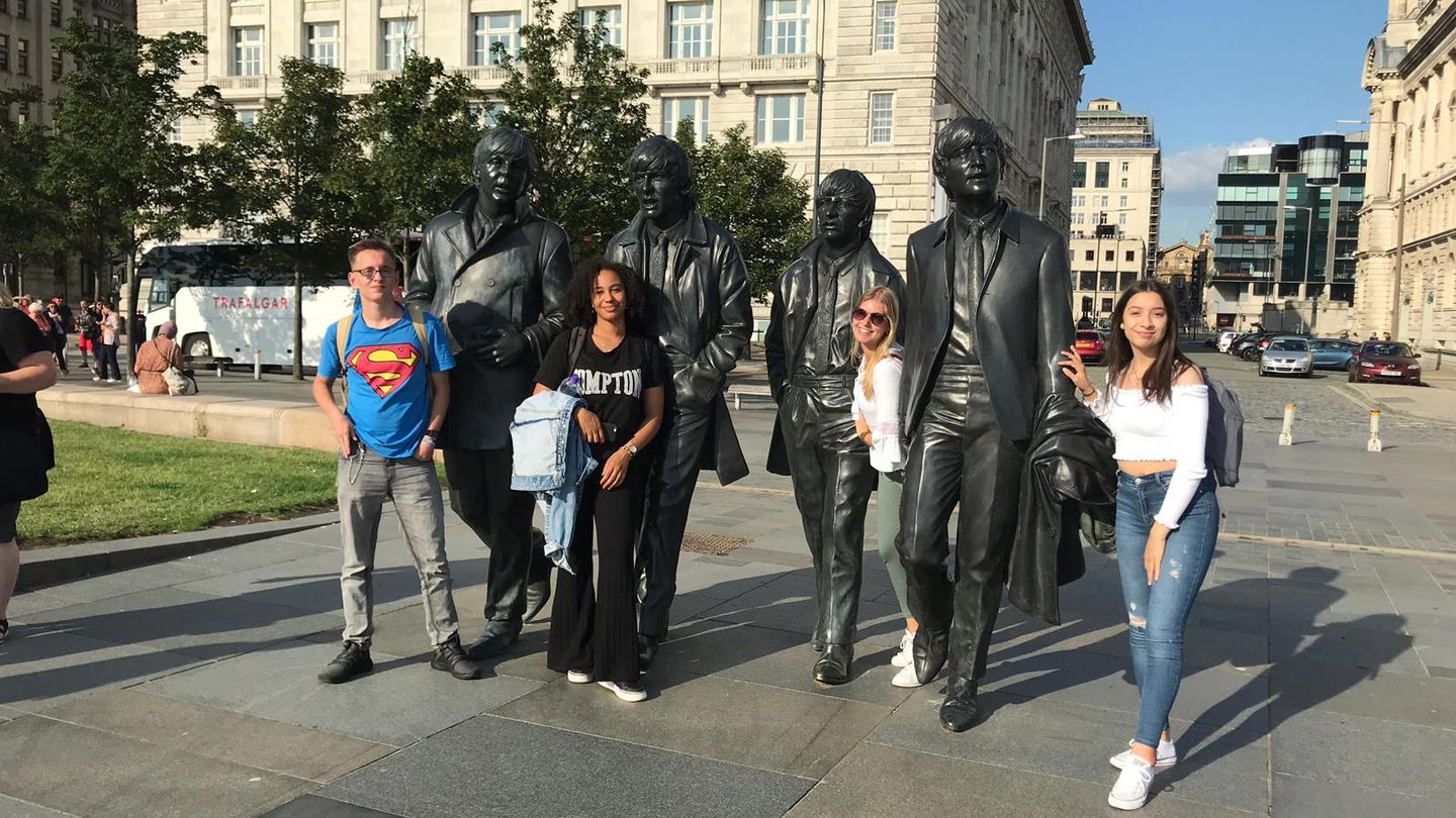 Vier Jugendliche, zum Teil mutmaßlich auch mit Migrationshintergrund, stehen zwischen überlebensgroßen Statuen der Beatles.