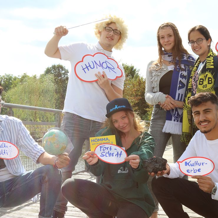 Eine Gruppe Jugendlicher steht oder hockt im Sonnenlicht auf einer Brücke. Sie halten Karten in die Höhe, auf denen z. B. "Fortschritt" oder "Humor" steht.