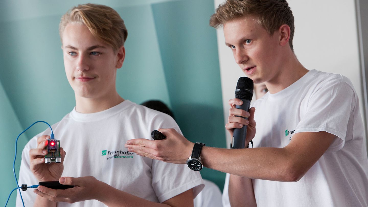 Zwei Jugendliche halten eine Präsentation. Der rechte spricht in ein Mikrofon, der linke hält ein elektronisches Bauteil in die Höhe, dessen Leuchte rot blinkt.