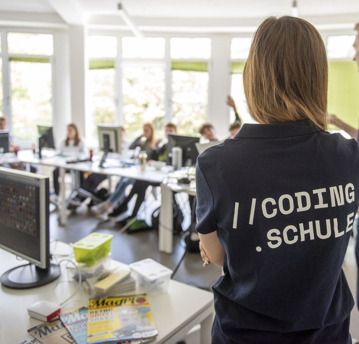 Im Vordergrund steht eine Person, die ein T-Shirt trägt, auf dem "Coding Schule" steht. Diese steht neben einem Schreibtisch mit Monitor, IT-Zeitschriften und weiteren Materialien. Im Hintergrund sitzen einige Jugendliche an Tischen, auf denen auch Monitore befestigt sind. Ein Jugendlicher hebt die Hand, um sich zu melden.