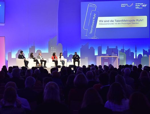 Eine große Veranstaltungshalle. Auf einer Bühne sitzen sechs Personen auf weißen Sesseln. Das Publikum ist in ein blaues Licht getaucht. Auf einer Leinwand ist u. a. zu lesen "Wir sind die TalentMetropole Ruhr".