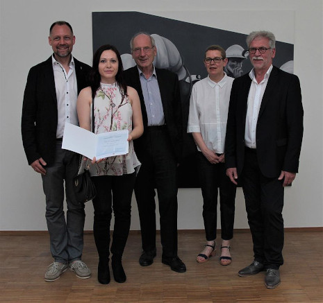 Gruppenbild der geförderten Stipendiatin und weiterer Projektbeteiligter vor einem Kunstwerk der Stipendiatin. Dieses zeigt Bergleute und ist in unterschiedlichen Grautönen gehalten. Die Stipendiatin hält eine Urkunde in die Höhe.