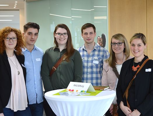 Geförderte Studierende stehen um einen Stehtisch mit weißer Husse herum und lächeln. Auf dem Stehtisch ist Schild, auf dem "RAG-Stiftung" steht.