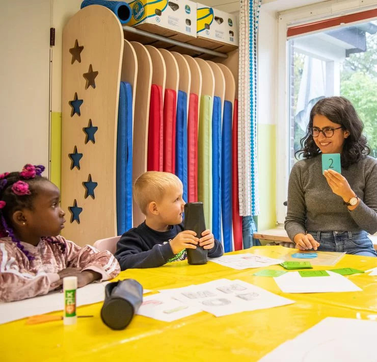 Eine Bildungsbegleiterin am Tisch mit zwei Kita-Kindern. Sie hält eine Karte mit der Ziffer "2" in die Höhe. Die Kinder sehen sie aufmerksam an.