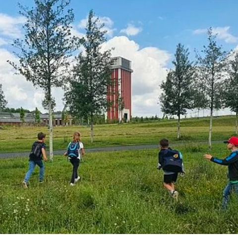 Vier Kinder mit Rucksäcken laufen über eine grüne Wiese. Im Hintergrund ist der Förderturm der Zeche Friedrich Heinrich zu sehen.