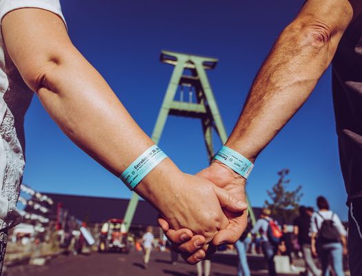 Zwei Personen halten sich an den Händen. Sie haben Armbänder an, die zur Einlasskontrolle dienen. Im Hintergrund ist ein Fördergerüst zu sehen. Der Himmel ist blau.