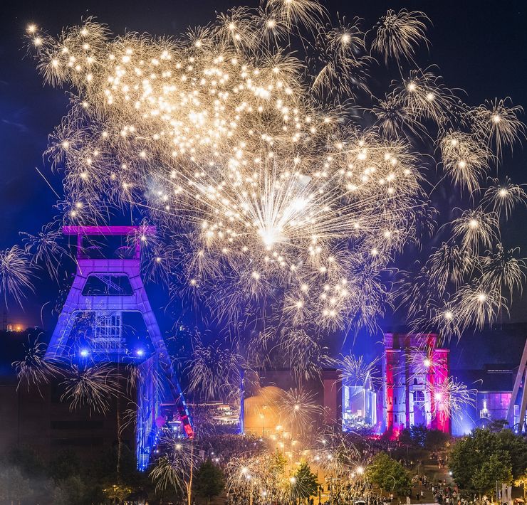 Zeche Zollverein in pink und blauen Farben angeleuchtet; am Nachthimmel ein spektakuläres Feuerwerk
