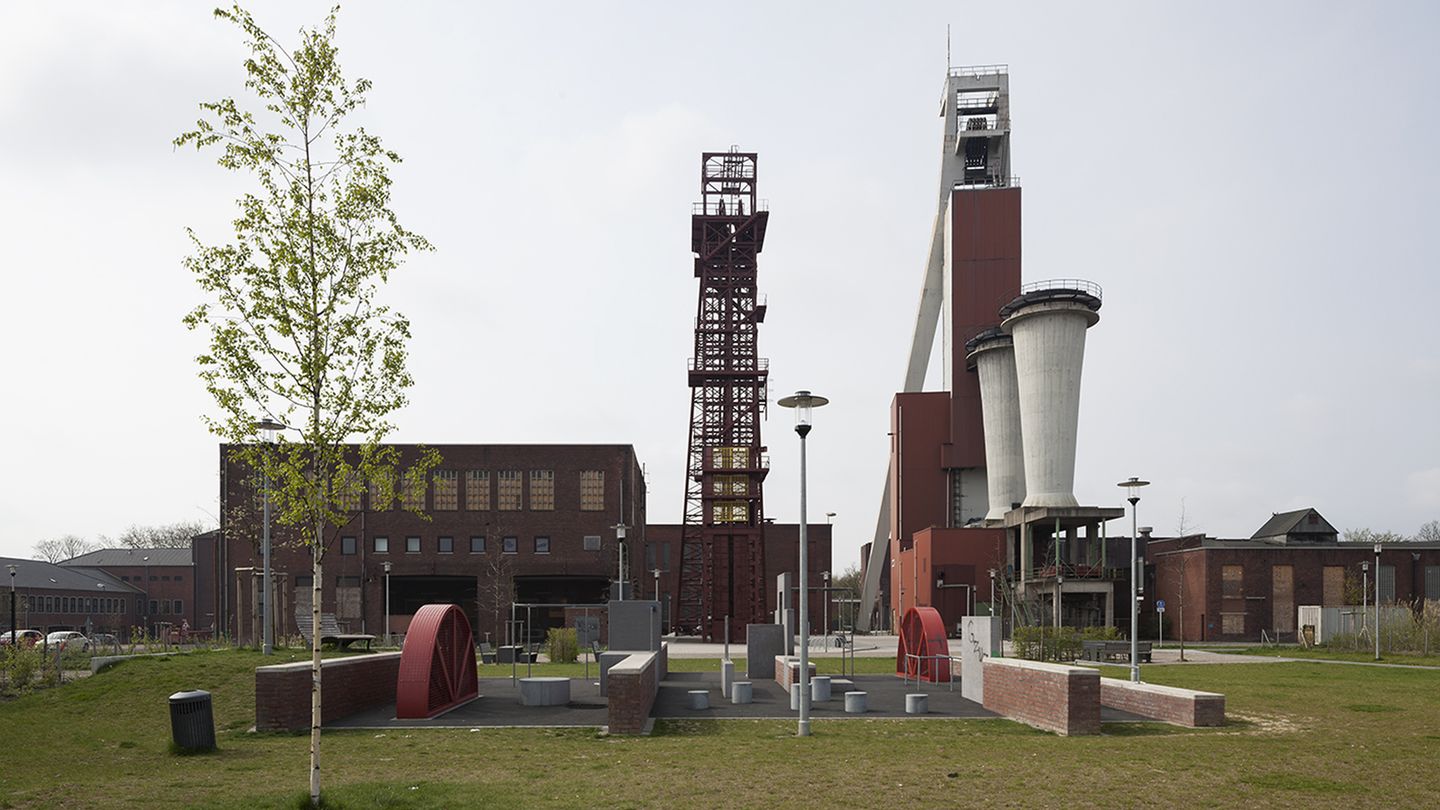 Links rot-braunes Backsteingebäude, mittig und rechts zwei Fördergerüste; im Vordergrund ein Aufenthaltsbereich