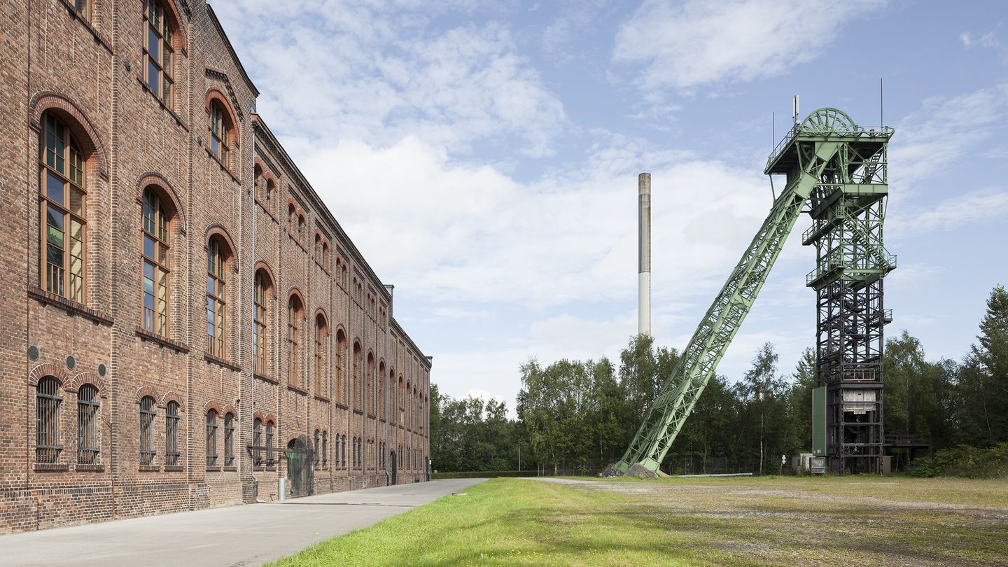 Auf der linken Seite sieht man ein braun-rotes Gebäude aus Backsteinen, die ehemalige Maschinenhalle, rechts ein Fördergerüst