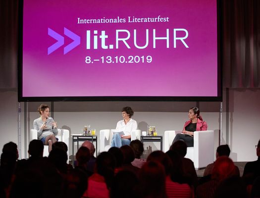 Drei Frauen auf einer Bühne vor Publikum. Im Hintergrund eine Leinwand, auf der "lit.RUHR" zu sehen ist 