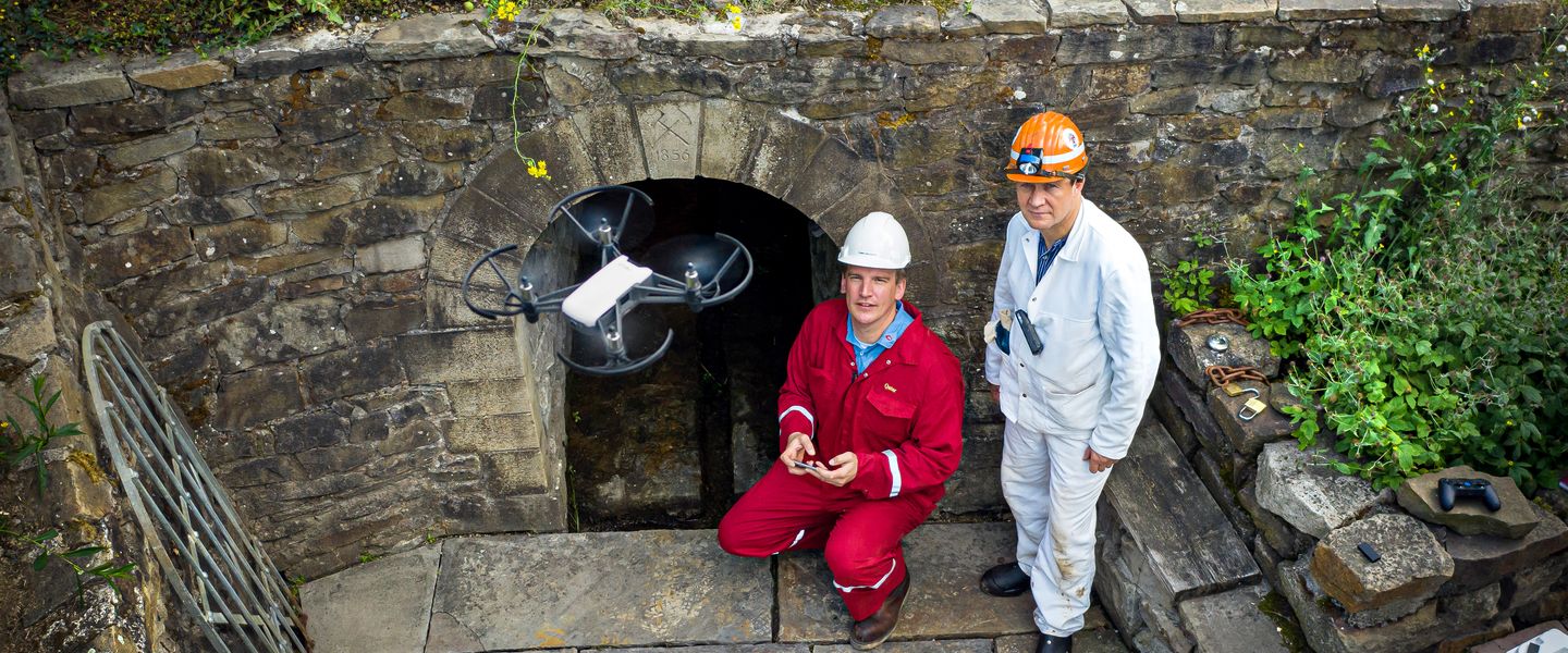 Zwei Wissenschaftler in Schutzkleidung und Helmen knien bzw. stehen vor einem niedrigen Tunneleingang. Der linke hält ein Steuerungselement in der Hand. Vor den beiden fliegt eine Drohne, die sich auf dem Weg in den Stollen befindet.