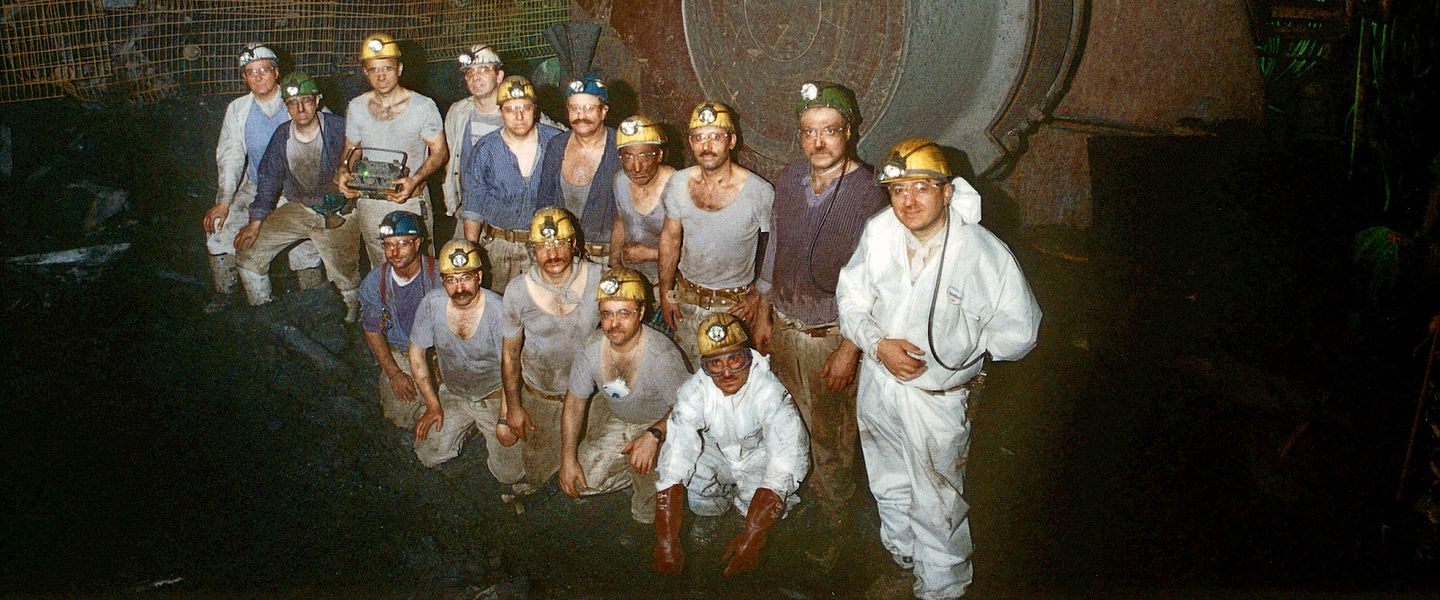 Gruppenfoto von Bergleuten unter Tage. Alle tragen Helme, Grubenlampen und sind schmutzig von Kohlenstaub