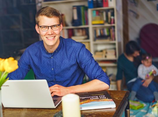 Ein Student mit Brille sitzt lächelnd an einem Schreibtisch vor einem Laptop. Neben ihm liegt ein Buch mit dem Titel "Die Welt der Rohstoffe". Im Hintergrund spielt eine Frau mit einem kleinen Kind auf ihrem Schoß.