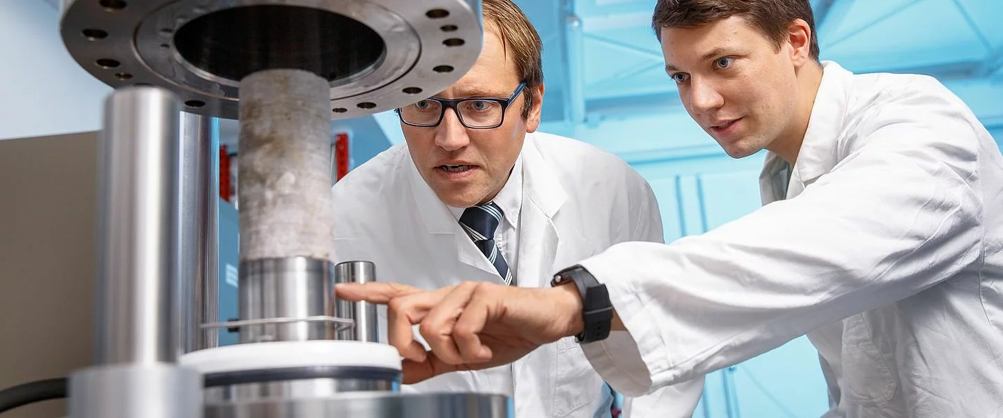 Zwei Wissenschaftler in Laborkitteln betrachten ein wissenschaftliches Messgerät. Der Jüngere zeigt mit seinem Finger auf das Gerät.