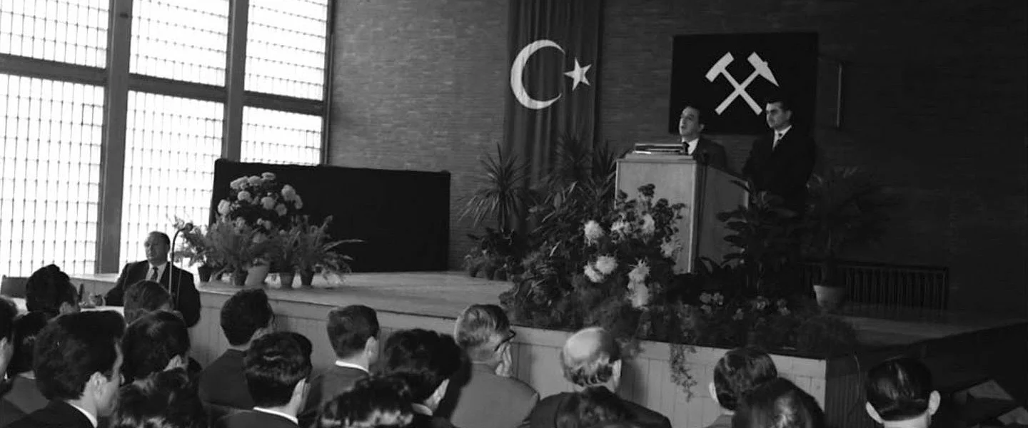 Schwarz-weiß-Foto, das eine Bühne steht hinter der die türkische Flagge sowie das Symbol des Bergbaus platziert sind. Zwei Personen stehen an einem Rednerpult, das mit Blumen geschmückt ist. Im Publikum sitzen viele Menschen, zum Teil mutmaßlich auch mit Migrationshintergrund
