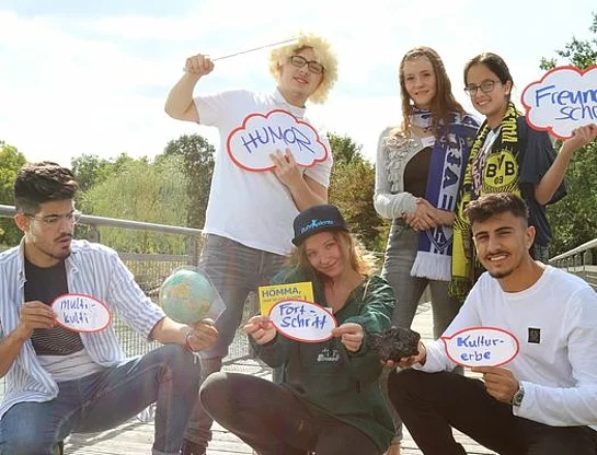 Eine Gruppe Jugendlicher steht oder hockt im Sonnenlicht auf einer Brücke. Sie halten Karten in die Höhe, auf denen z. B. "Fortschritt" oder "Humor" steht.